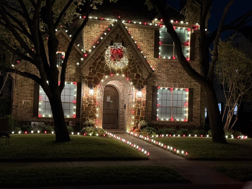 Front view of a brick Dallas home decorated with warm white and red Christmas lights outlining the roofline and windows, with pathway lighting leading to the entryway.